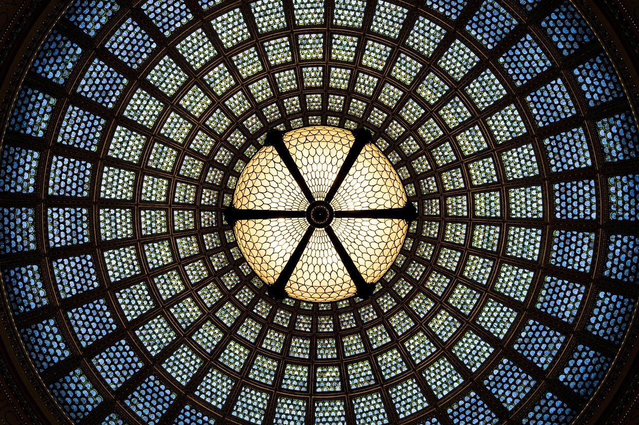 Intricate stained glass dome viewed from below, featuring a geometric pattern of circles and hexagons in shades of blue, green, and yellow. The design radiates from a central point, creating a symmetrical and ornate appearance.
